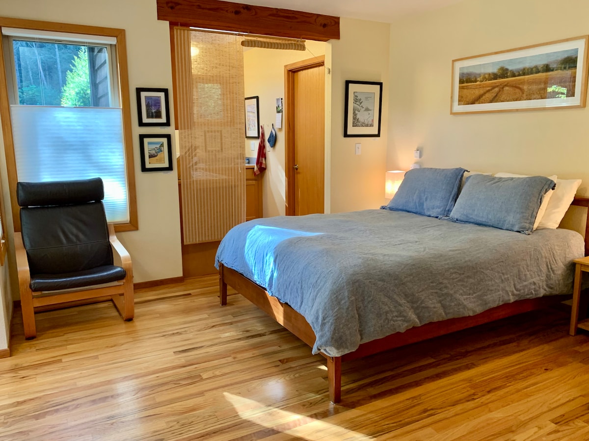 A spacious sleeping area features a queen-sized bed with soft blue bedding. The warm hardwood floors complement the wooden furniture. A comfortable chair and framed artwork are visible, while natural light filters through the window, illuminating the inviting room.