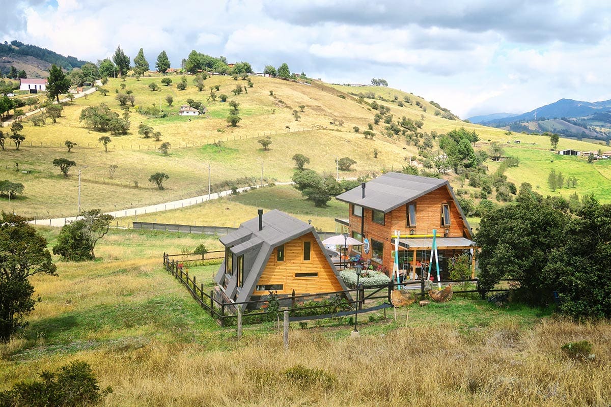 A serene landscape is captured, featuring two wooden houses nestled in a green and golden hillside. A fenced yard surrounds the houses, and gentle rolling hills are visible in the background, complemented by a cloudy sky.