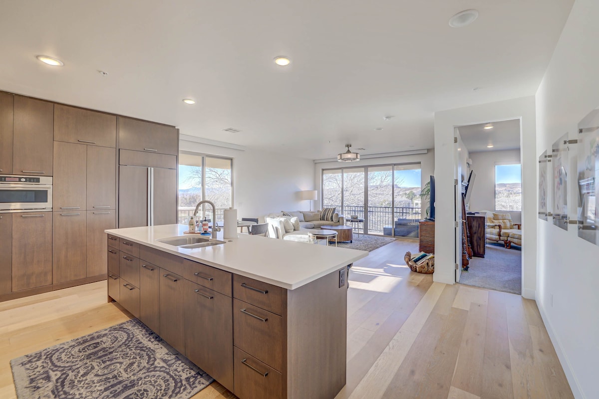 A modern kitchen features sleek wood cabinetry and a spacious island with a sink. Open to the living area, large glass doors provide access to a balcony, allowing natural light to fill the space. A cozy seating area and neutral decor enhance the welcoming ambiance.