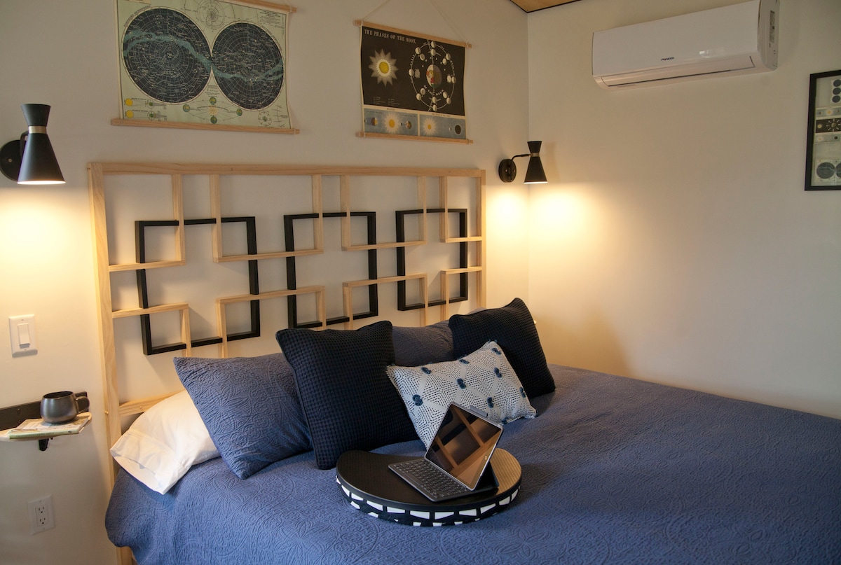 A cozy bedroom features a modern wooden headboard with a geometric design. A soft blue quilt covers the spacious bed, accented by several dark-patterned pillows. A small tray holds a laptop on the bed, while wall-mounted lamps provide soft lighting. Astronomical prints adorn the walls.