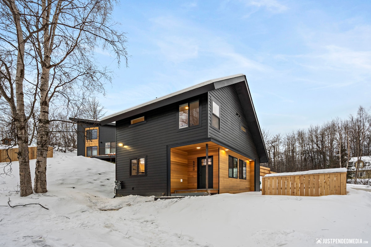 A modern two-story home is depicted, featuring a sleek black and wood exterior. Snow blankets the surrounding ground, enhancing the structure's contemporary design. Large windows provide natural light, and a welcoming entryway leads into the residence.