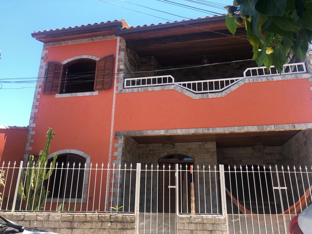 The exterior of a two-story house features vibrant orange walls and stone accents. Wooden shutters frame the windows, and a white fence encloses the front yard, which includes a large cactus. A covered balcony is visible above the entrance, providing additional outdoor space.