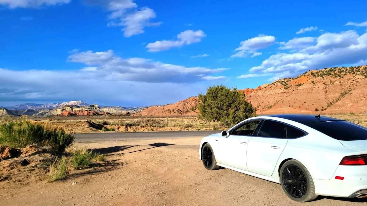 A white car is parked on a gravel surface, with a scenic view of rolling hills and distant mountains in the background. The sky is mostly clear with scattered clouds, and the landscape features natural earthy tones complemented by patches of green vegetation.