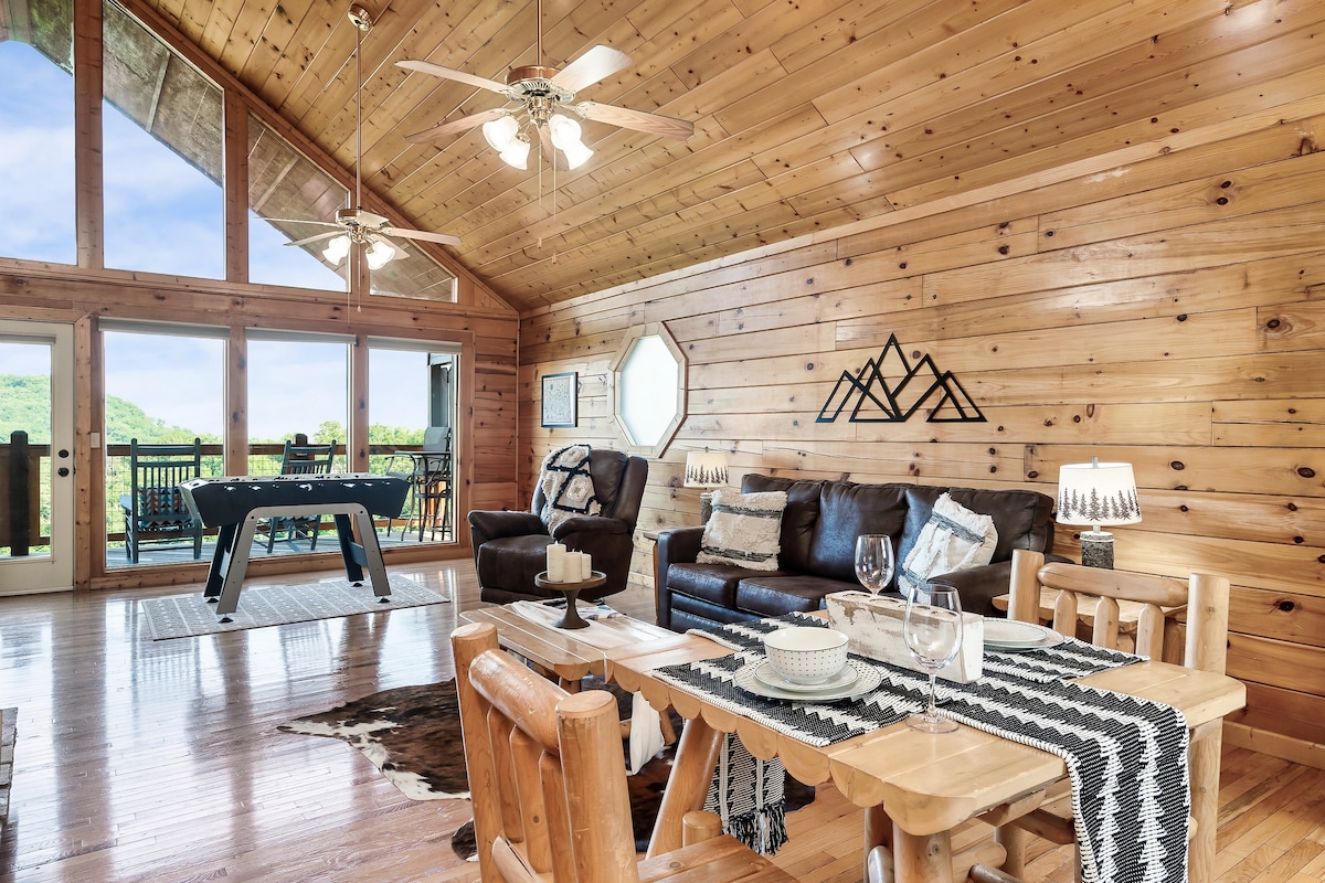 The spacious Great Room features a comfortable leather couch and wooden dining set, with a foosball table visible in the background. Large windows allow natural light to flood the space, showcasing views of the surrounding mountains. Wood paneling creates a rustic ambiance.
