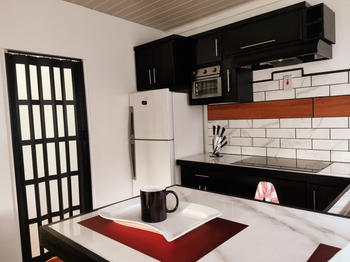 A modern kitchen showcases sleek black cabinetry and white marble countertops. A white plate is placed on the table, accompanied by a matching black mug. A stainless steel refrigerator stands prominently, while a glass door allows light into the space.