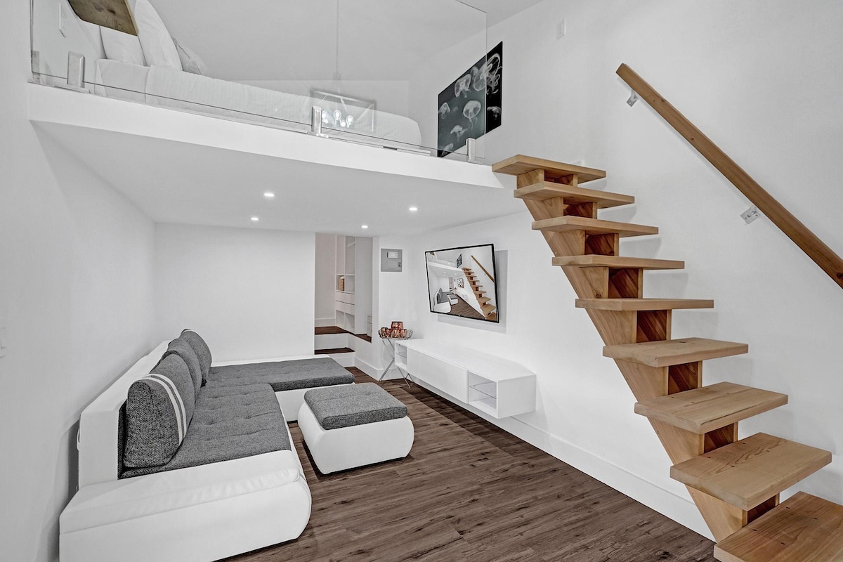 A stylish loft interior is visible, featuring a seating area with a modern sectional sofa and a minimalist entertainment unit. Light wood stairs lead to an upstairs bedroom with a queen-size bed, illuminated by soft ceiling lights.