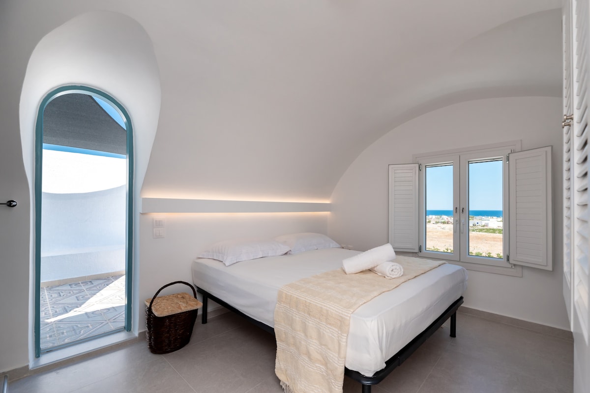 A bright and airy bedroom features a spacious bed with crisp white linens. A woven basket is placed beside the bed. Natural light enters through a large window and a rounded door that leads to outdoor space, creating a serene atmosphere.