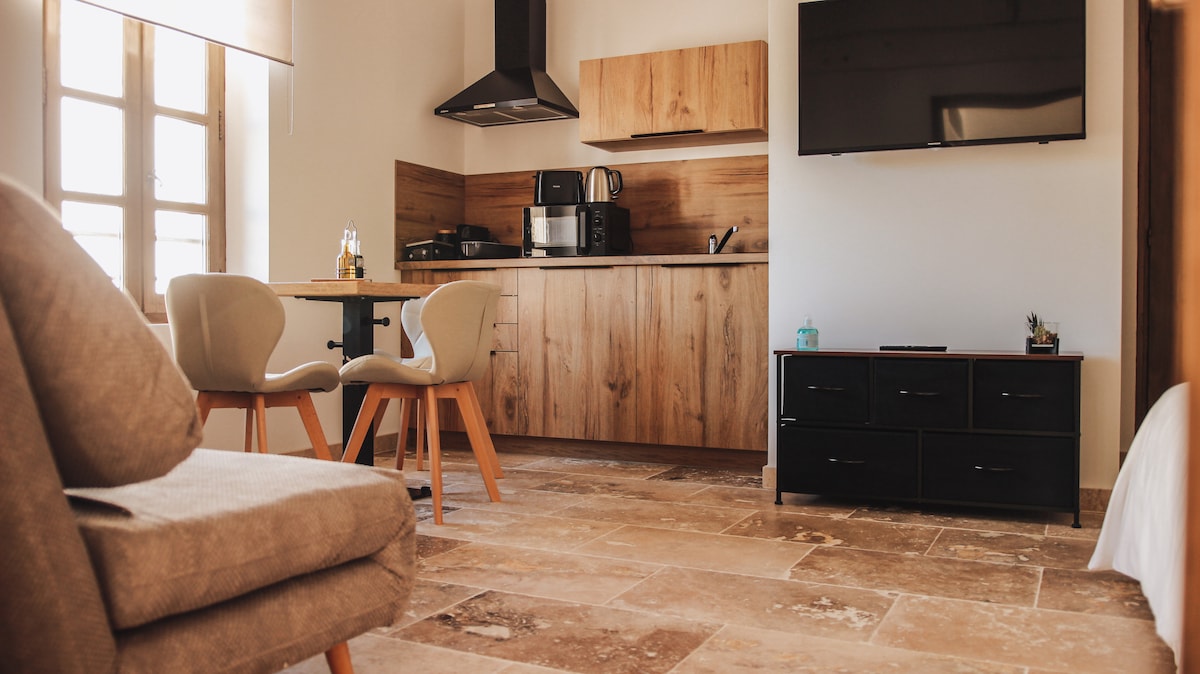 A modern kitchen area is situated alongside a dining table with two chairs, featuring warm wooden tones. Nearby, a comfortable sofa offers a relaxed seating option. A flat-screen television is mounted on the wall, complementing the overall functional layout of the space.