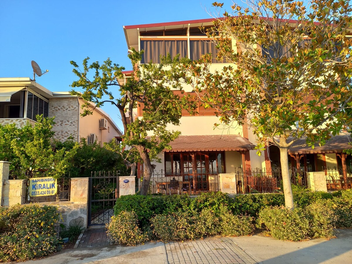 The exterior of a two-story building is visible, surrounded by lush greenery and flowering shrubs. A shaded outdoor seating area is featured under a wooden awning, with a sign indicating rental availability. The bright blue sky enhances the welcoming appearance of the structure.