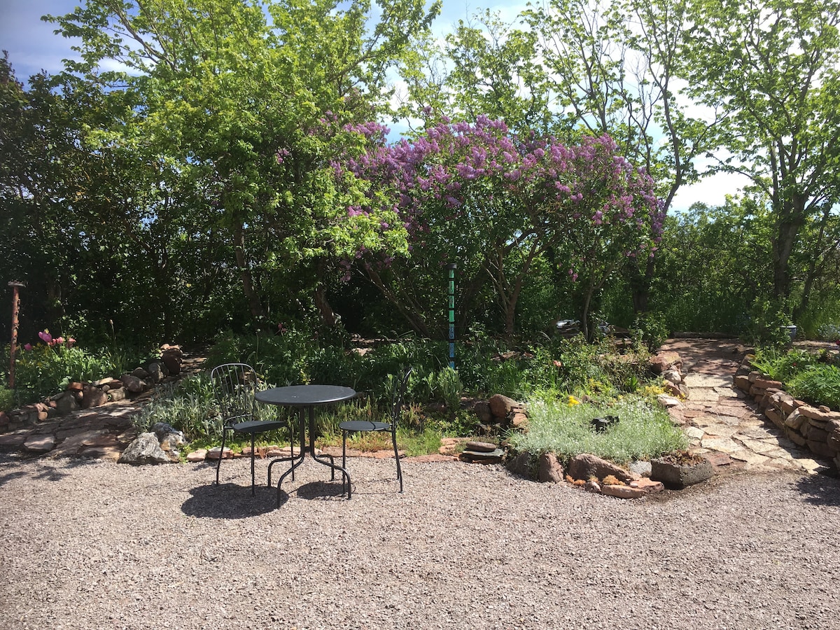 An outdoor seating area features a small round table accompanied by two black chairs, set on gravel. Lush greenery surrounds the space, including flowering plants and trees, creating a serene garden environment. A stone pathway is visible leading through the garden.