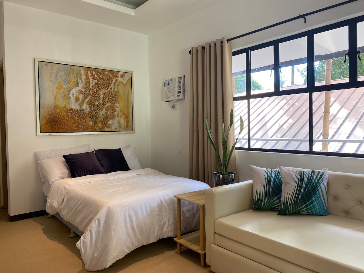 A bright sleeping area features a spacious bed with crisp white bedding and contrasting dark pillows. A light-colored sofa with decorative cushions is positioned nearby. Large windows allow natural light to stream in, with views of greenery outside.