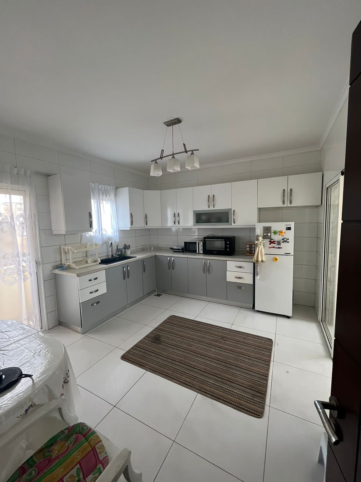 The kitchen is equipped with modern appliances, including a microwave and refrigerator. Light-colored cabinetry contrasts with sleek gray accents. A small rug is positioned on the tiled floor, and natural light enters through a nearby window, creating a bright and functional cooking space.
