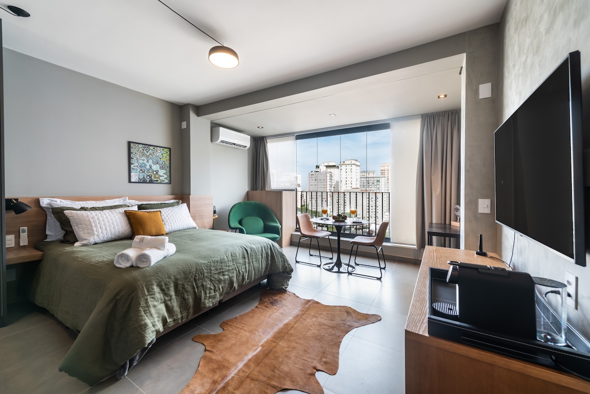 A modern studio apartment features a spacious bed adorned with soft linens and decorative pillows. A bright area with large windows provides a view of the city. A dining table for two sits beside a comfortable armchair, with a smart TV positioned on the side.