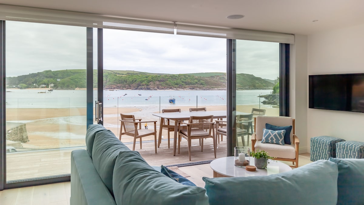 A spacious living area features large glass sliding doors opening to a balcony, offering views of the beach and sea. Soft teal furnishings provide comfort, while a round coffee table and a light accent chair complete the inviting ambiance. A smart TV is mounted on the wall.