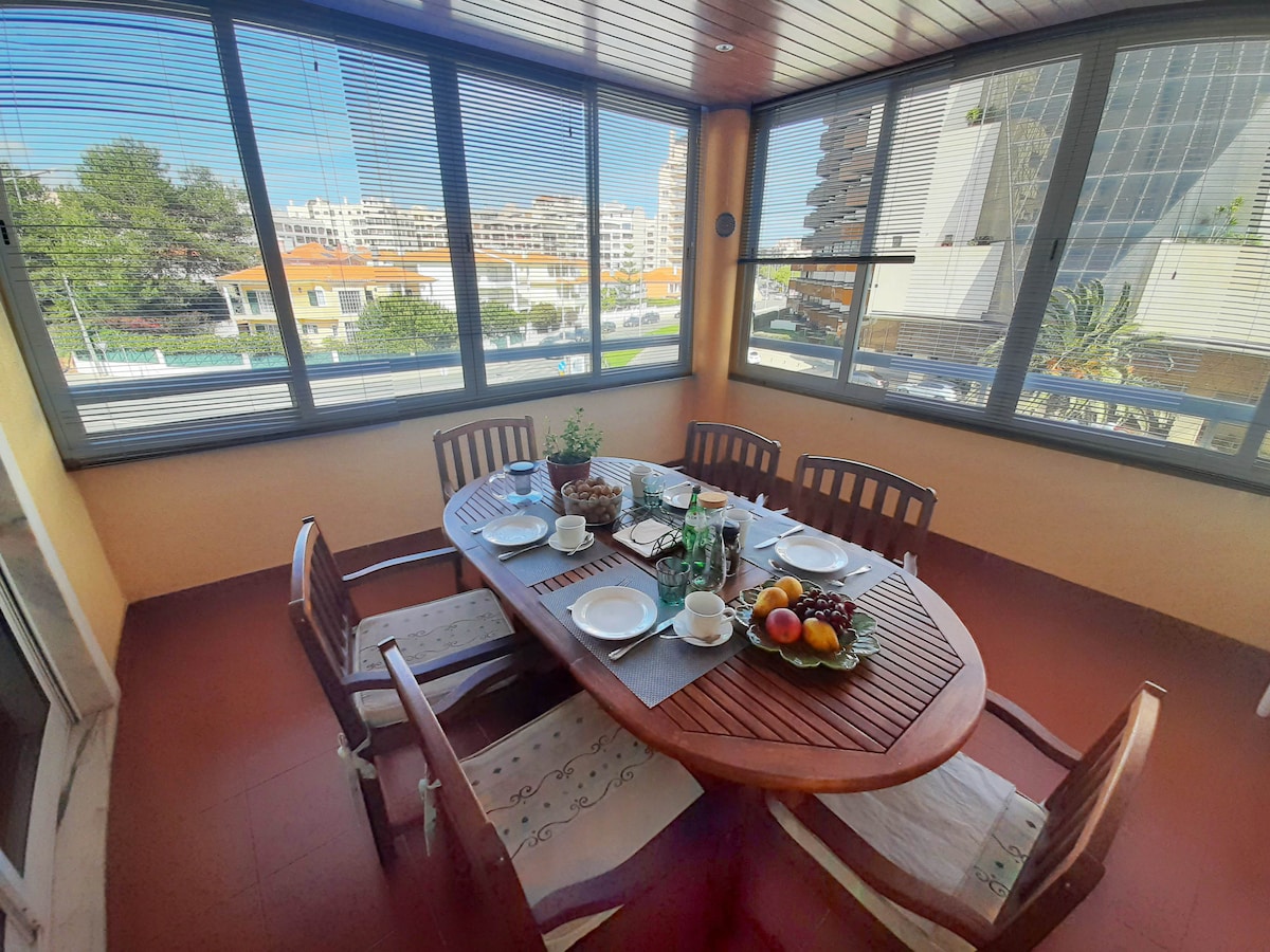A dining area with an oval wooden table is shown, surrounded by six wooden chairs. The table is set with plates, cutlery, and a fruit centerpiece. Large windows allow for natural light and provide views of the surrounding area.