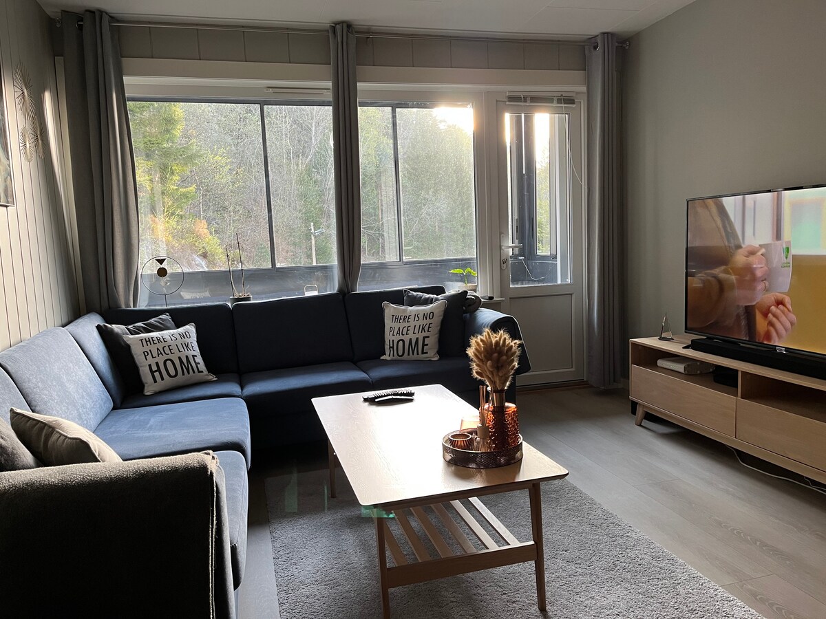 A spacious living area features a large sectional sofa with decorative pillows. Natural light floods in through multiple windows, illuminating a coffee table and an entertainment unit with a television. Soft rugs are placed on light-colored flooring, creating a comfortable atmosphere.