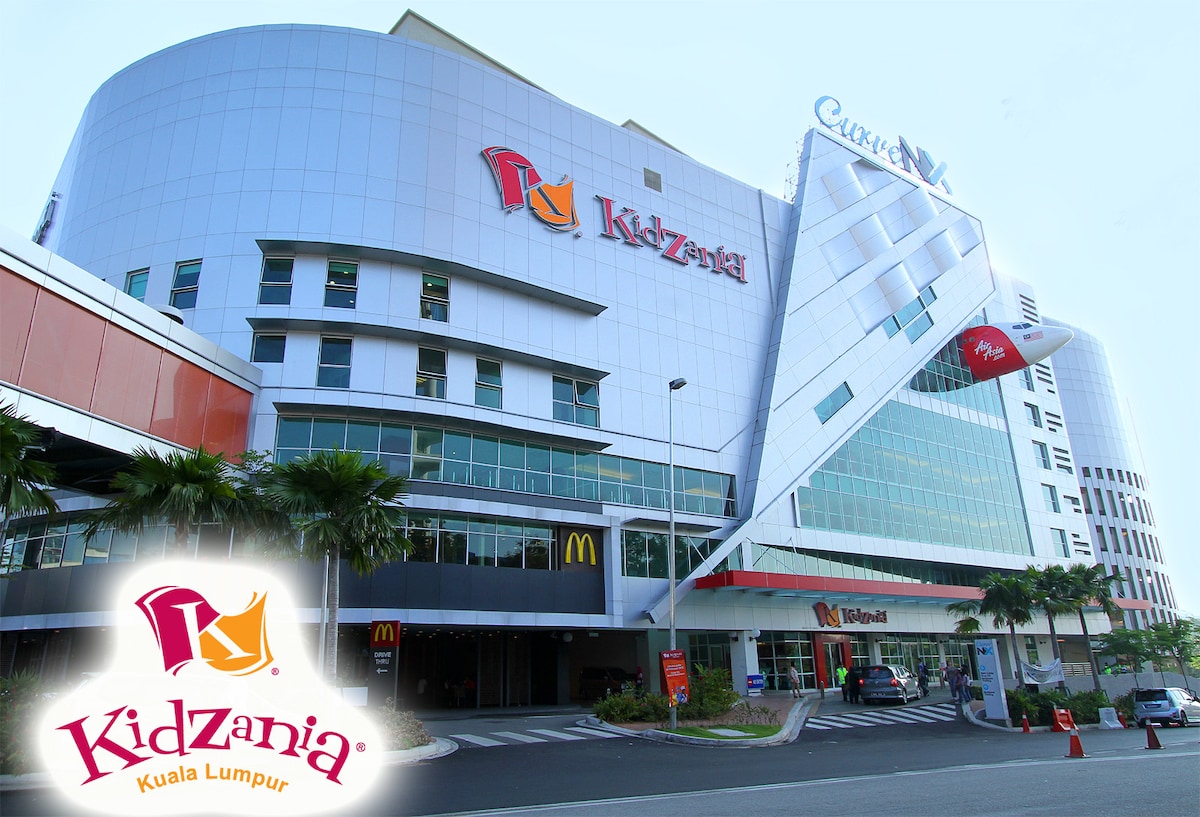 The image displays the exterior of KidZania Kuala Lumpur, showcasing its modern architecture. A prominent red and yellow logo is visible, accompanied by a McDonald's outlet at the entrance. Palm trees are positioned nearby, enhancing the vibrant urban atmosphere.