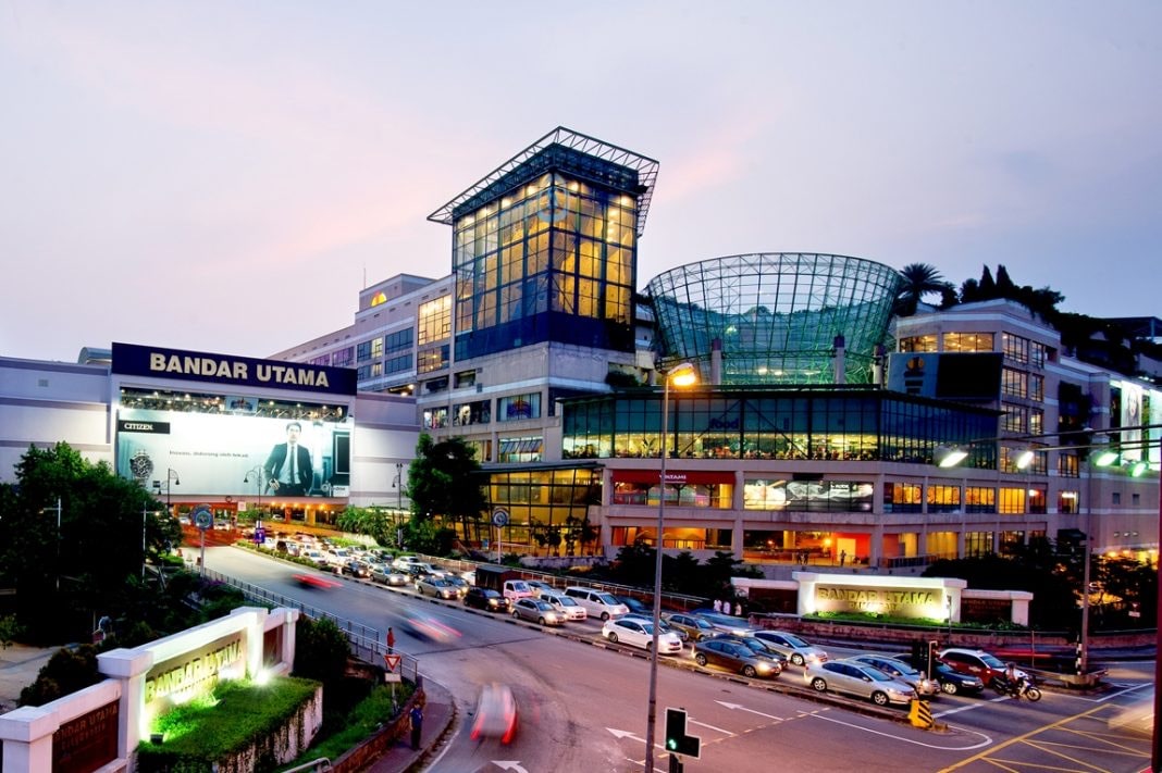 A modern shopping mall is featured with multiple glass-paneled structures, showcasing a blend of retail and dining spaces. Surrounding the building, a bustling street lined with vehicles is visible, and the evening sky casts a colorful backdrop to the lively scene.