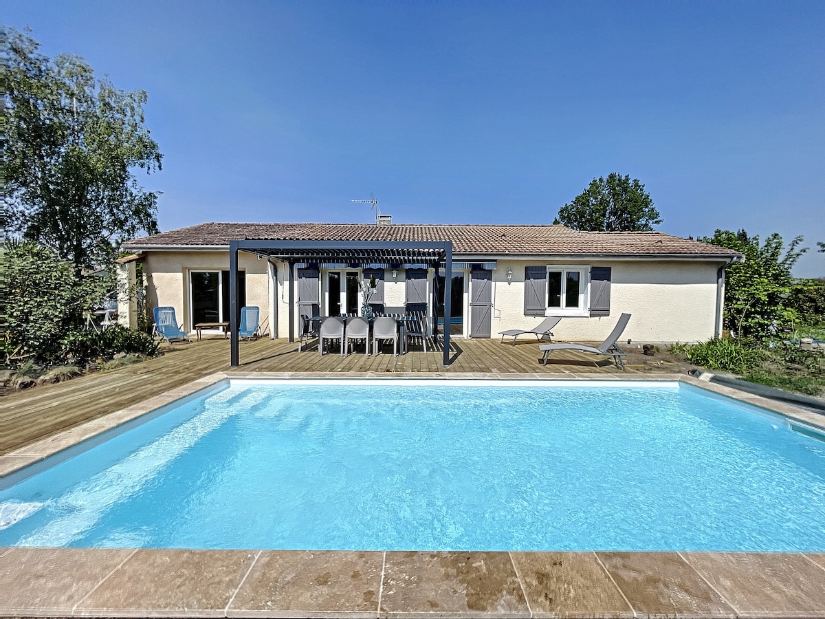 A modern house with a wooden terrace is shown next to a heated swimming pool. Sun loungers and dining seating are arranged under a pergola, providing a shaded area for relaxation. Lush greenery surrounds the property, enhancing its tranquil environment.