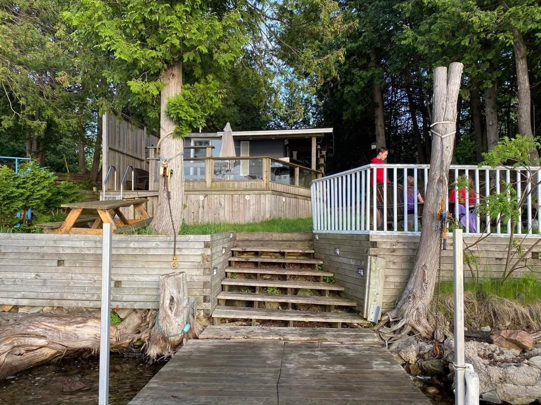 A pathway of wooden steps leads from the water's edge to an elevated deck area. The cottage is visible in the background, surrounded by trees. Seating areas, including a picnic table, are positioned nearby, creating a welcoming outdoor space for relaxation.