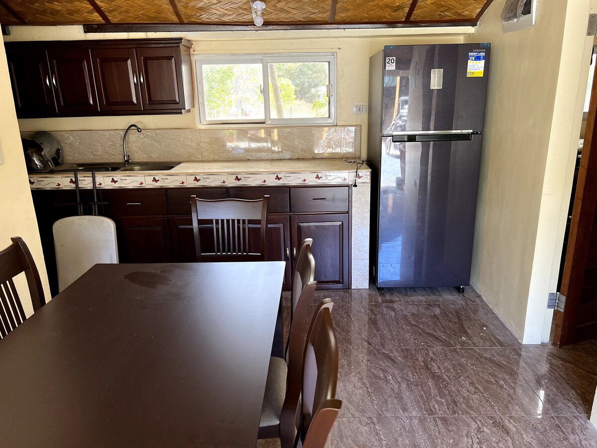 A well-equipped kitchen area features dark wooden cabinetry and a large refrigerator. A dining table with six chairs occupies the space, complemented by a sink and countertop for meal preparation. Natural light enters through a window, illuminating the clean, tiled flooring.