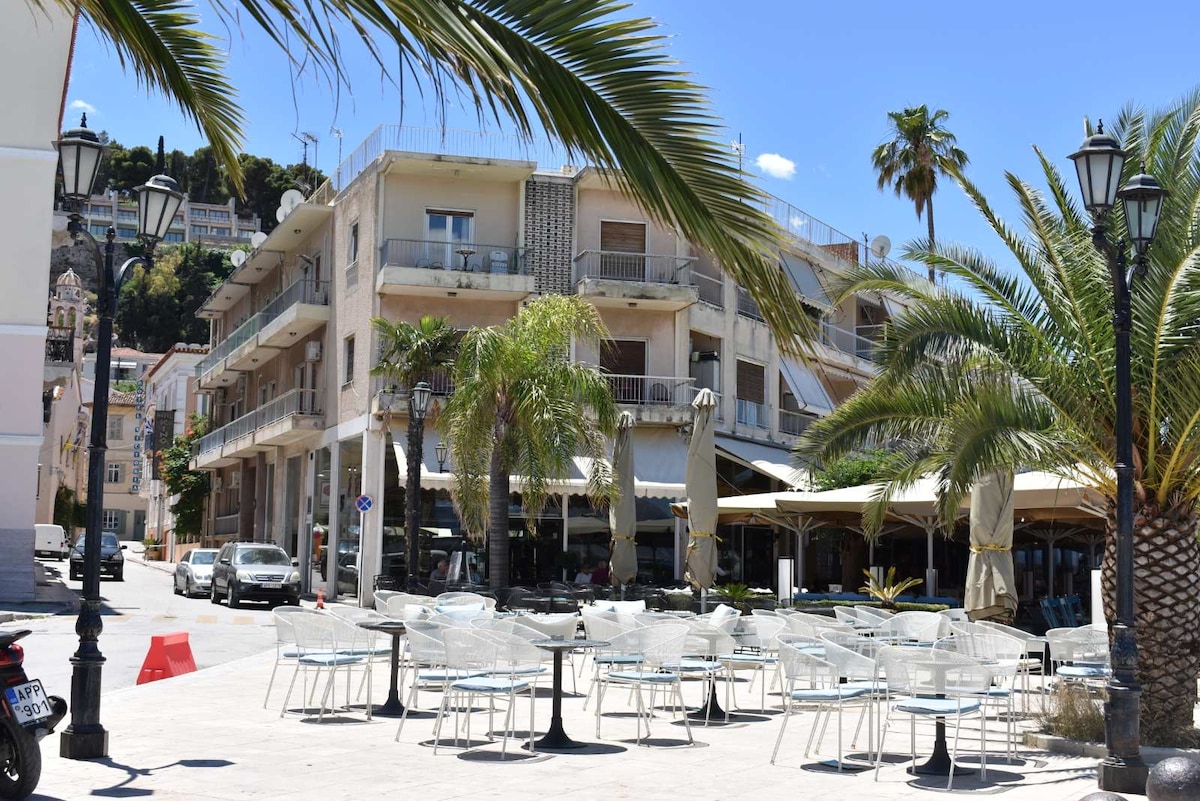 An outdoor seating area is shown with transparent chairs and round tables arranged on a sunlit square. Nearby palm trees and buildings are visible, with a clear blue sky above. The scene captures a vibrant atmosphere close to the historic center.