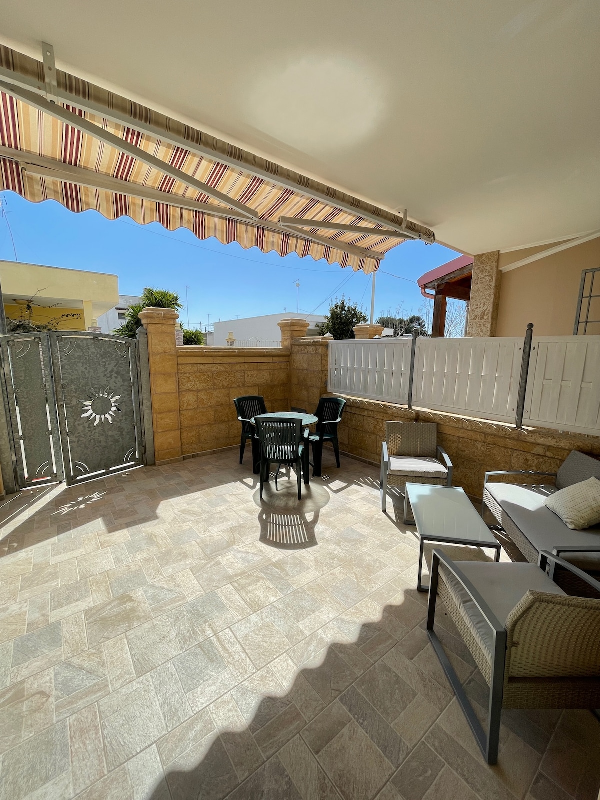 An outdoor seating area is displayed, featuring several black and light-colored chairs arranged around a small table. A striped awning provides shade overhead while light filters in, illuminating the tiled floor. Stone walls and a decorative gate add privacy to the space.
