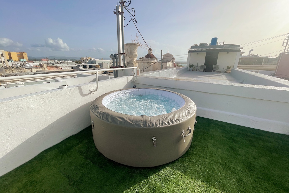 A circular jacuzzi is featured on the rooftop, surrounded by lush green artificial grass. The area overlooks the city and offers a clear view of the horizon and distant buildings under a bright sky.