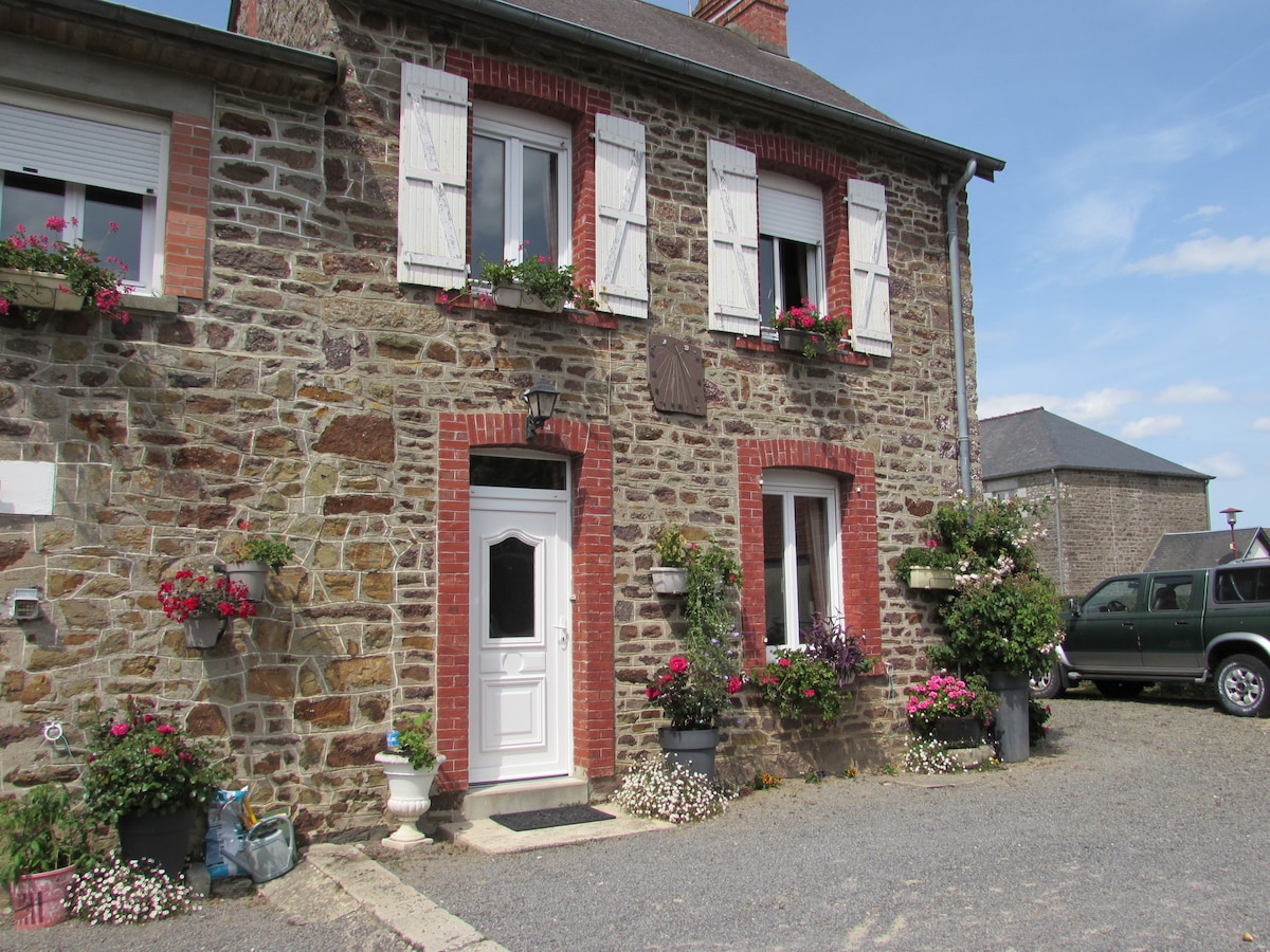 A rustic stone house with red brick accents is presented, featuring white shutters and flower boxes filled with colorful blooms. A welcoming front door is framed by a garden, while gravel creates a natural path leading to the entrance.