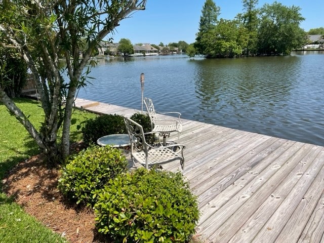 A serene lakefront deck is shown with two decorative chairs and a small table, surrounded by lush greenery. The calm water reflects the trees and nearby homes, creating a peaceful environment for relaxation.