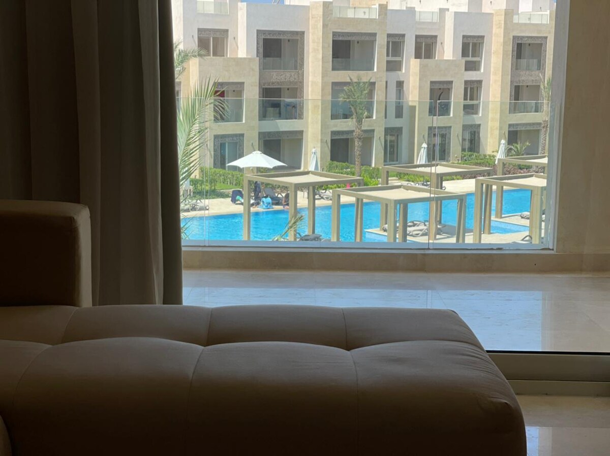 A view from inside the apartment showcases a spacious outdoor pool area, bordered by palm trees and sun loungers. Bright sunlight illuminates the scene, highlighting the relaxing environment. The large window frames the tranquil setting, offering a glimpse of the vibrant pool water.