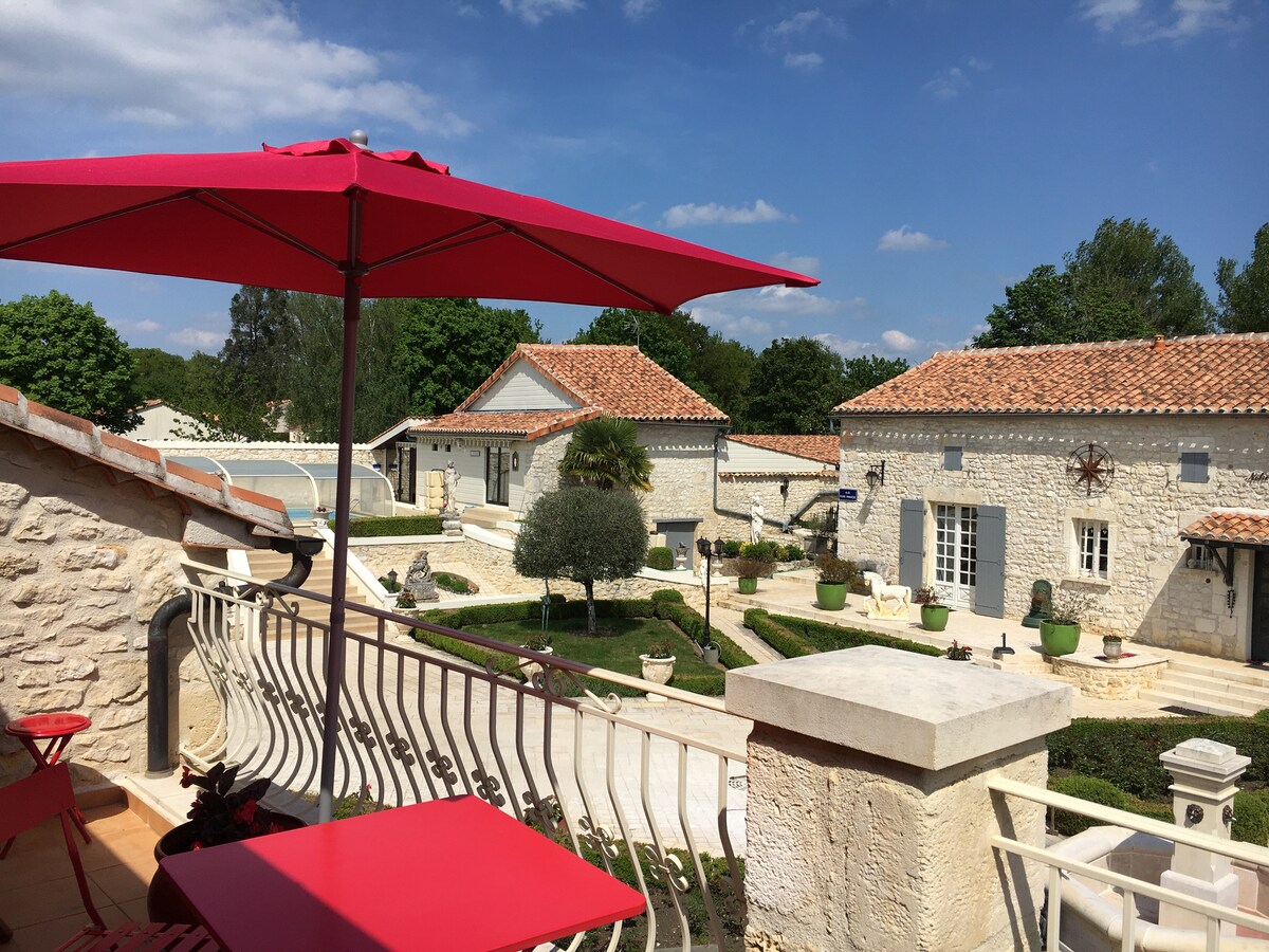 A terrace with a red umbrella provides a view of the landscaped courtyard, featuring stone buildings with traditional roofs. Lush greenery and decorative planters create a serene outdoor setting, while the blue sky enhances the tranquil atmosphere.