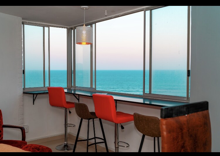 A seating area features a counter with two high chairs, offering unobstructed views of the ocean through expansive windows. The space is brightened by a stylish pendant light, and the sky showcases soft pastel hues blending into the water.