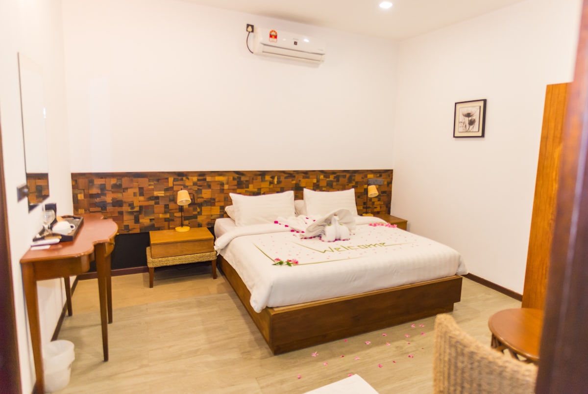 A cozy bedroom is equipped with a spacious bed adorned with decorative pillows and towel art. Bedside lamps provide soft lighting beside the bed. A wooden table and wicker chair are positioned near the door, while a mirror reflects the room's design.