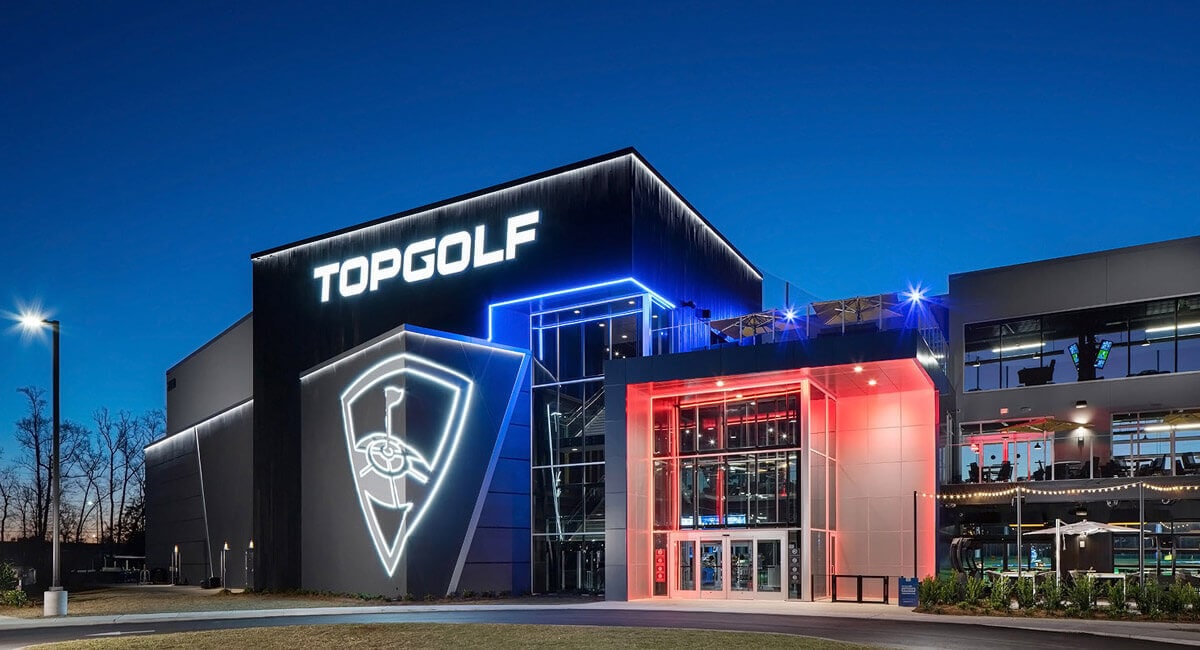 The modern Topgolf venue is illuminated against the evening sky, featuring distinct neon lighting. The exterior showcases large glass windows and a prominent logo, highlighting the vibrant entertainment space. The surrounding area is well-lit, emphasizing accessibility for visitors.