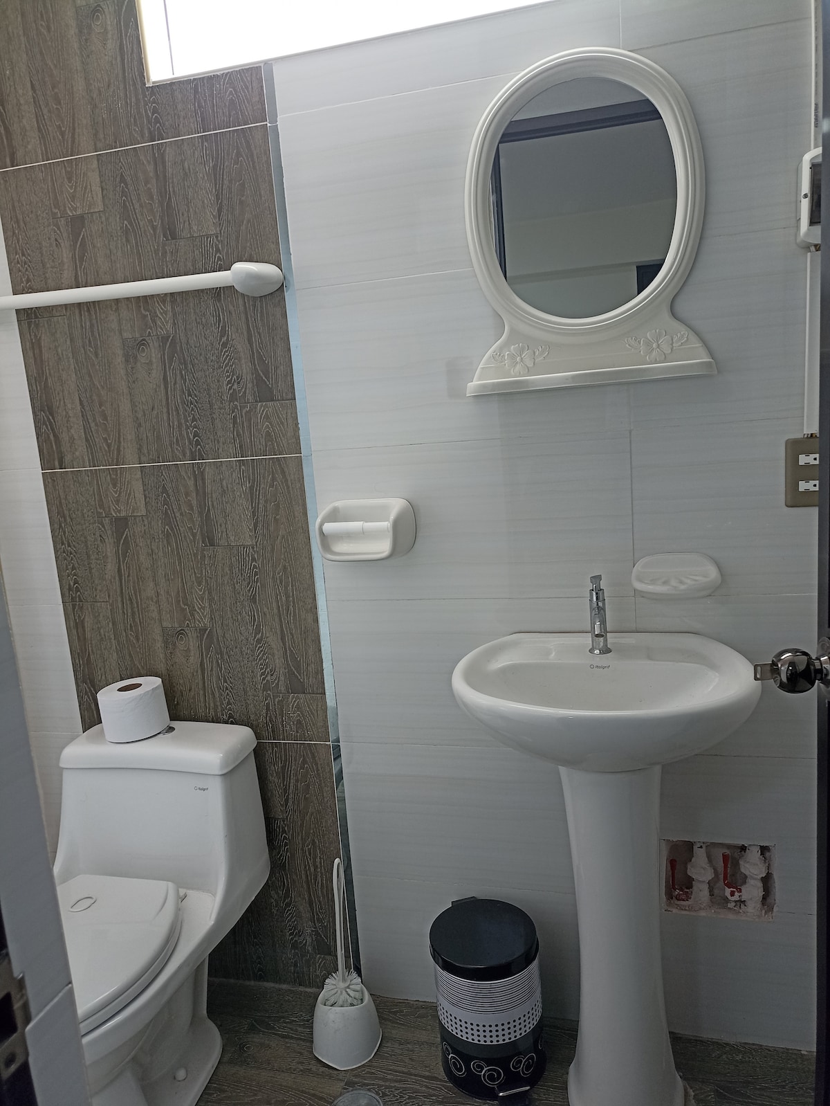 A compact bathroom is shown, featuring a white sink with a modern faucet positioned above a sleek pedestal. A clean toilet is visible, accompanied by a small toilet paper holder. An ornate mirror reflects light near the window, enhancing the space's brightness.