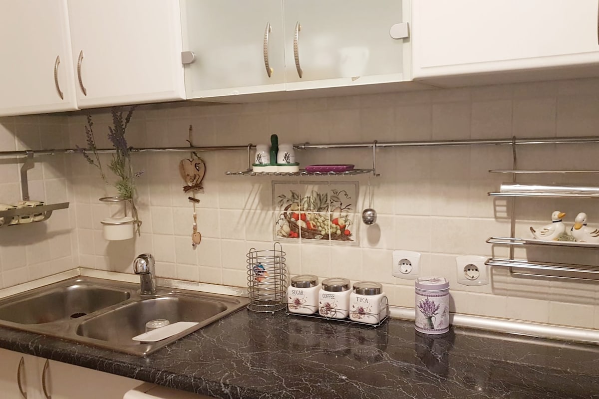 The kitchen features a black countertop with a stainless steel sink. Wall-mounted shelves hold decorative containers and kitchen utensils. The white cabinetry provides a clean and organized look, while subtle decorative accents, like a lavender motif, add charm to the space.