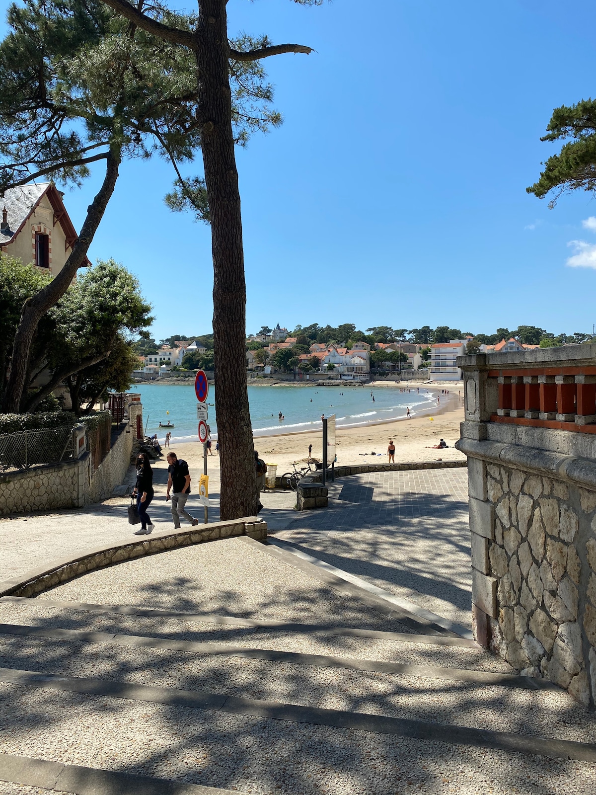 A view down a stone pathway lined with trees, leading toward a sandy beach. The shoreline is visible in the distance, with people enjoying the area and boats on the water. Nearby, a residential area is discernible, showcasing a mix of buildings among the greenery.