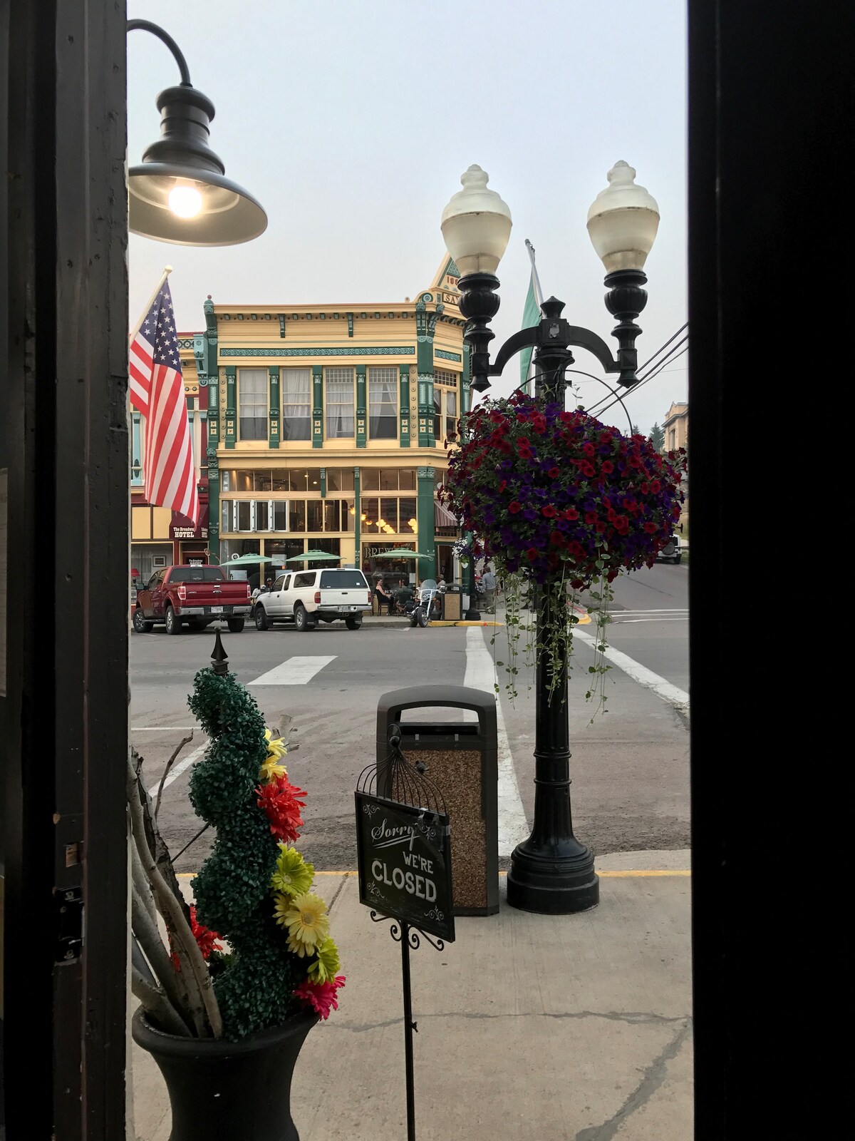 A view from the doorway showcases the vibrant downtown area, featuring an American flag and a historic building in the background. Flower baskets hang from a nearby lamppost, while a closed sign and decorative planters frame the entrance, creating an inviting street scene.