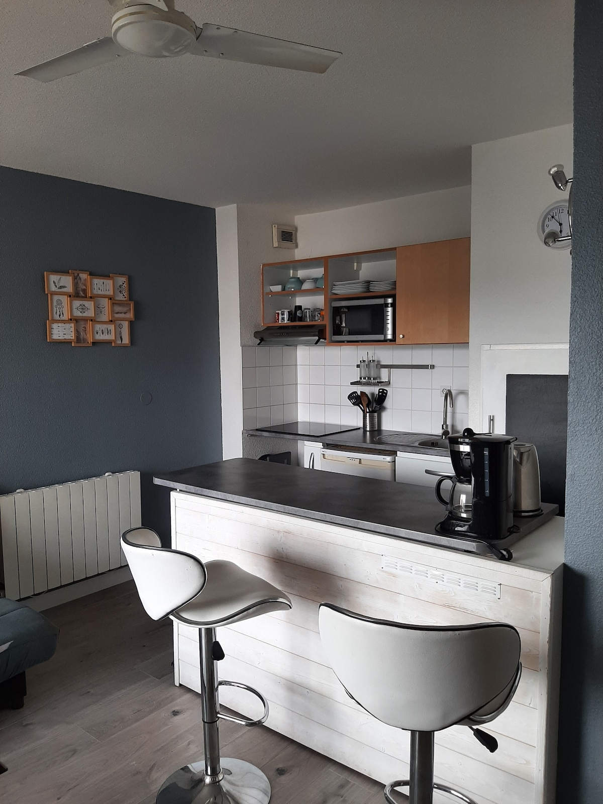 A compact kitchen area features modern appliances, including a microwave and coffee maker. A stylish bar counter with two white stools invites casual dining, while sleek cabinetry offers ample storage. A ceiling fan enhances ventilation, and a framed arrangement of artwork decorates the nearby wall.