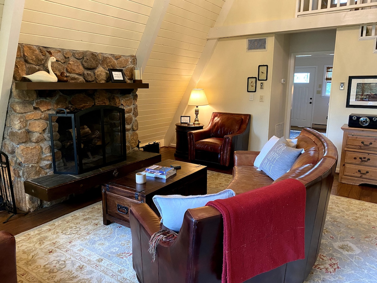 A warm living area features a stone fireplace as the focal point, flanked by two leather chairs and a cozy sectional sofa adorned with a red throw. A coffee table sits in front, and natural light enters through large windows, highlighting wood details.