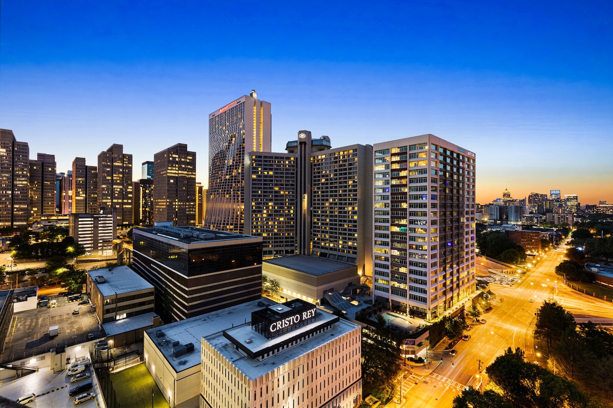 The city skyline is captured during twilight, revealing illuminated buildings and a vibrant atmosphere. Streets below are lined with lights, showcasing the lively urban scene. The penthouse location offers sweeping views of this stunning backdrop, enhancing the appeal of the top-floor condominium.