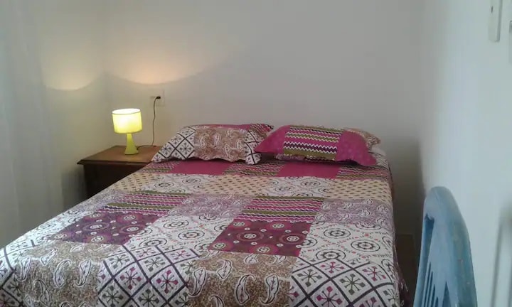 A comfortable bedroom features a double bed adorned with a colorful patchwork quilt and matching pillows. A small bedside table with a yellow lamp is present, providing warm light. The room is bright, with a neutral wall color that enhances the cozy feel.