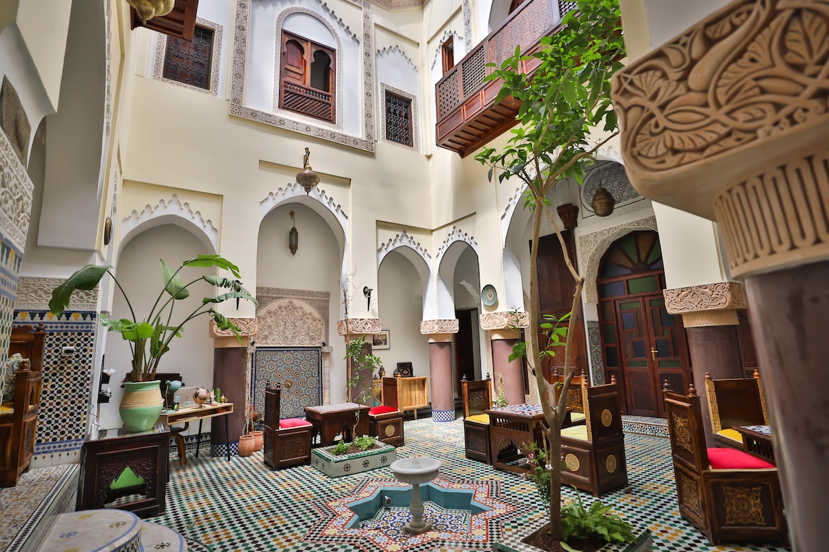 An interior courtyard features intricately tiled flooring and a calming fountain at the center. Surrounding the fountain, wooden seating with colorful cushions is arranged beneath ornate arches. Lush plants and traditional Moroccan decor enhance the serene atmosphere, providing a glimpse into the craftsmanship of the riad.