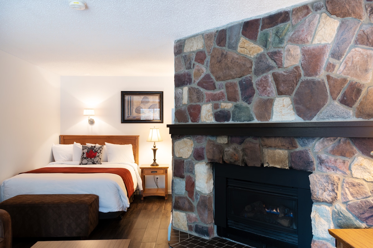 A cozy room features a spacious bed with decorative pillows, accented by a warm-toned comforter. A stone fireplace with a gas insert serves as a focal point. Soft lighting is provided by a bedside lamp, creating a comfortable ambiance for relaxation.