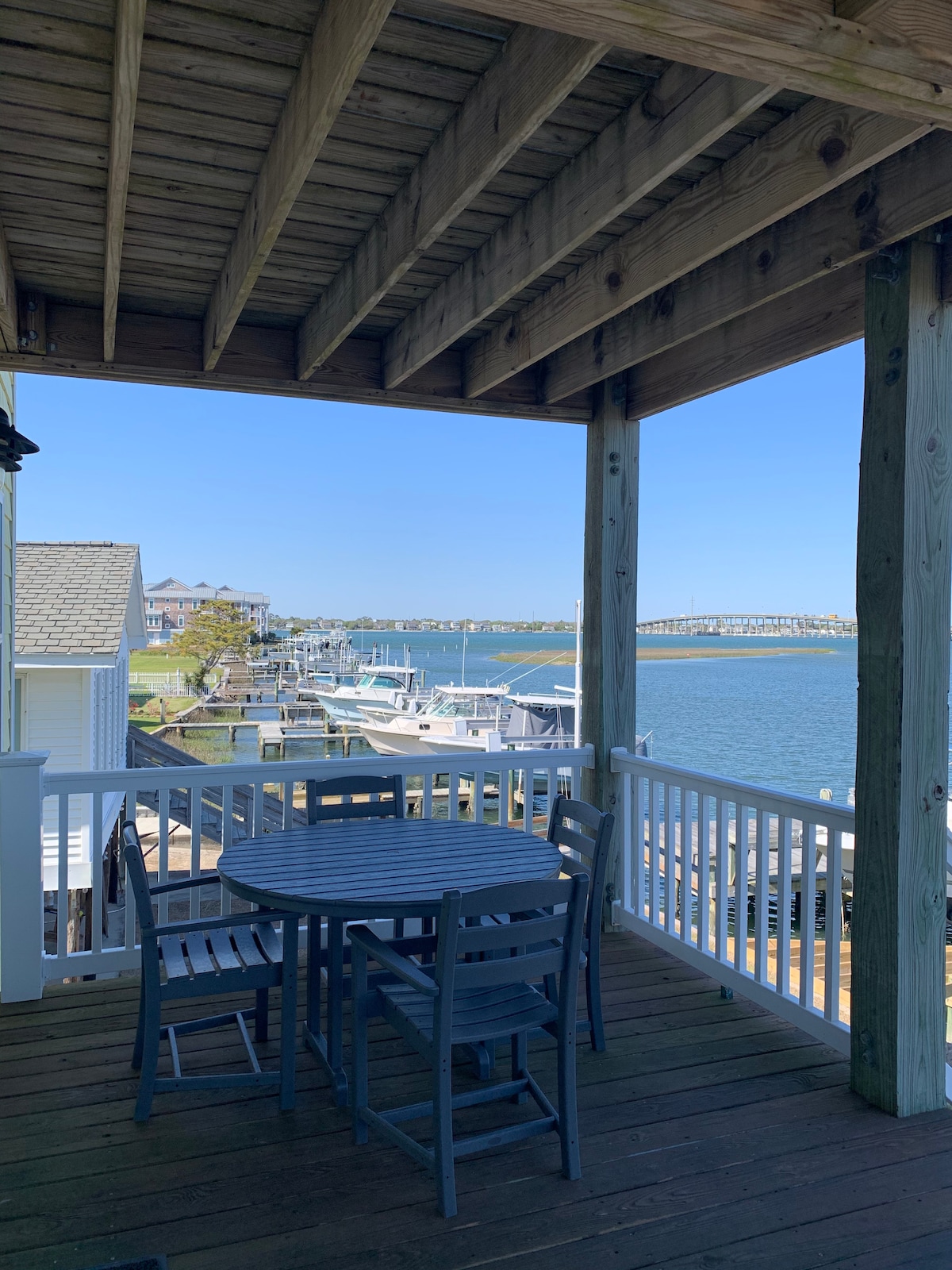 A shaded porch area features a table surrounded by four chairs, overlooking the tranquil waters of Moonlight Bay. Views of nearby docks and boats are visible, while the AB Bridge spans across the horizon under a clear blue sky.