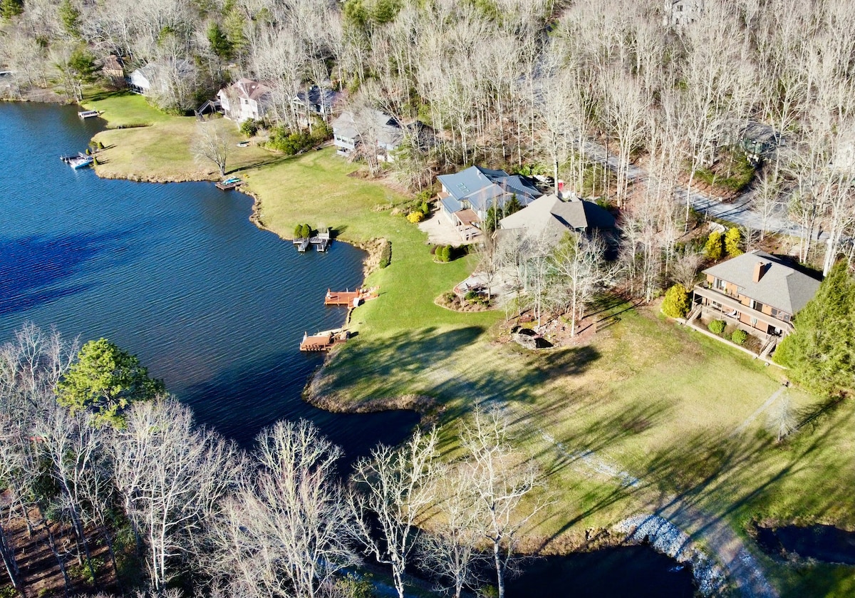 An aerial view showcases a serene lakeside setting with several well-maintained properties nestled among trees. The lush green lawn extends to the water's edge, and private docks are visible along the shoreline, inviting outdoor activities.