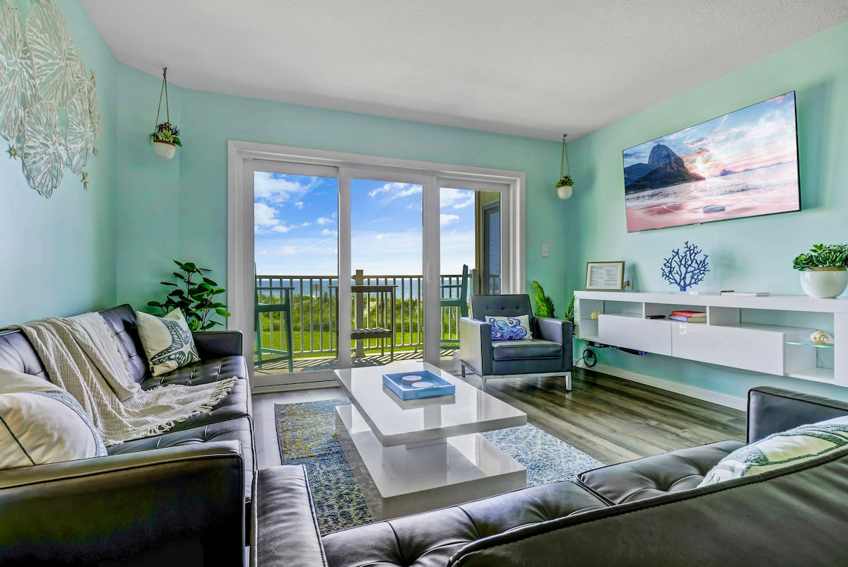 The living room features large sliding doors that open to a private balcony, providing a view of the outdoors. Comfortable seating is arranged around a coffee table, with greenery enhancing the space. A smart TV is mounted above a modern console, adding to the contemporary decor.