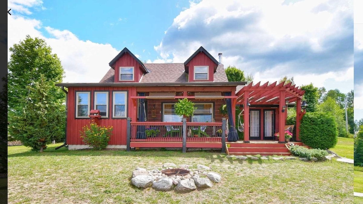 A charming red cabin features a welcoming front porch with a wooden pergola. Lush greenery surrounds the property, while a stone fire pit is positioned in the front yard, providing a perfect space for outdoor relaxation. Large windows allow natural light to fill the interior.