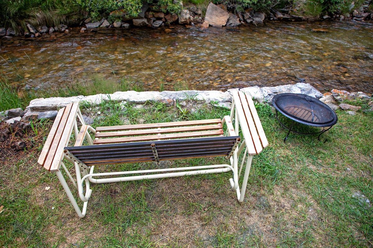 A simple wooden bench is positioned by the edge of a clear creek, providing a space to relax and enjoy the sound of flowing water. A fire pit sits nearby, surrounded by grass and stone, ideal for gatherings outdoors.